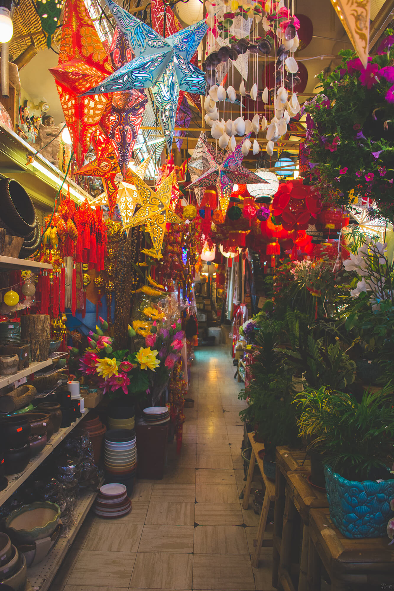 An aisle in Bamboo Village. The shelves that hug the wall on the left are lined with flowerpots and household ceramics, whiel the bamboo stools along the right side of the aisle display plants of various sizes. Low-hung star-shaped lanterns and windchimes made of seashells guide the way down the narrow corridor of goods and handicrafts.