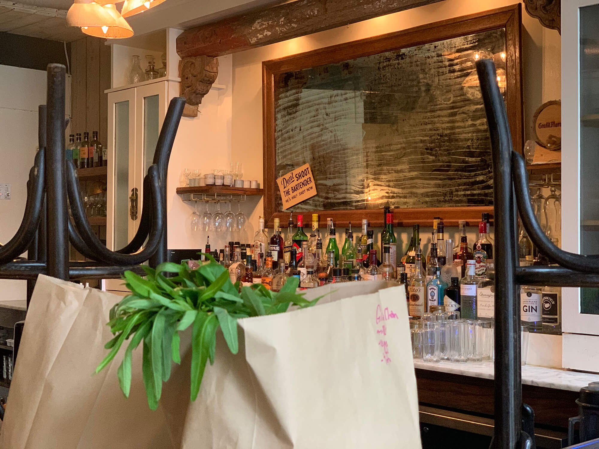 View of the bar. Liquor bottles of various colours and sizes are lined in tiers along the wall, above this display is a large tarnished mirror with a sign on the bottom left that reads 'Don't shoot the bartender, he's half shot already'. In the foreground are two paper bags with groceries. Leafy green vegetables peek out from on the of bags.