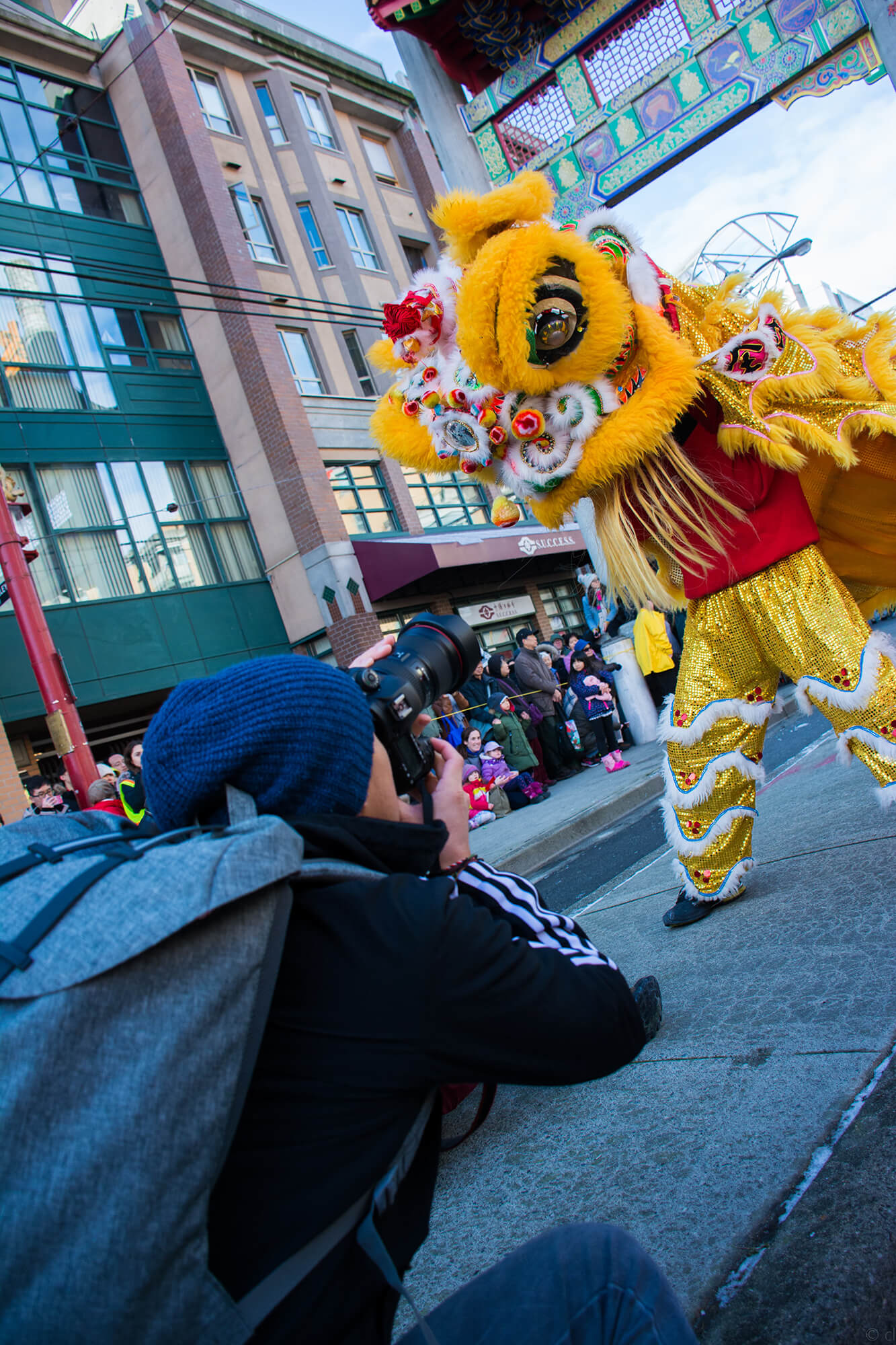 A photographer is captured laying down in prone position on the sidewalk with residual snow between its cracks, capturing a snapshot of the raised lion head during a parade performance from below. 正所谓，螳螂捕蝉，黄雀在后.