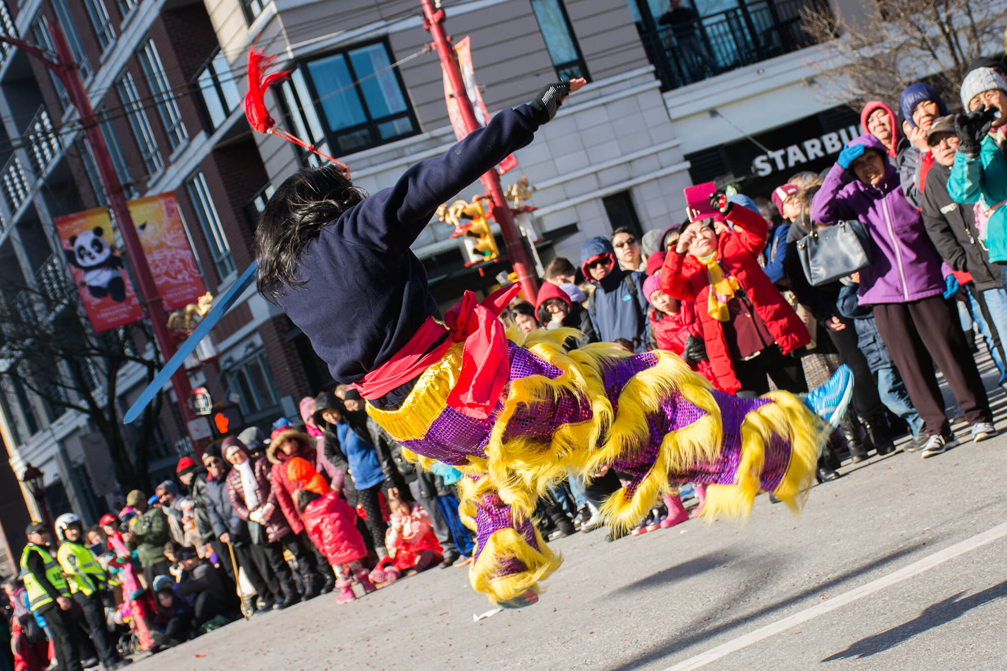 A member of the Chau Luen Athletic Club performs a sword set for audiences in a parade. The performer is mid-jump, twisting away from the camera. They hold a long sword in their left hand and move with such force that the tassles attached to the handle are shot straight up in the air as the performer lands from the jump. 