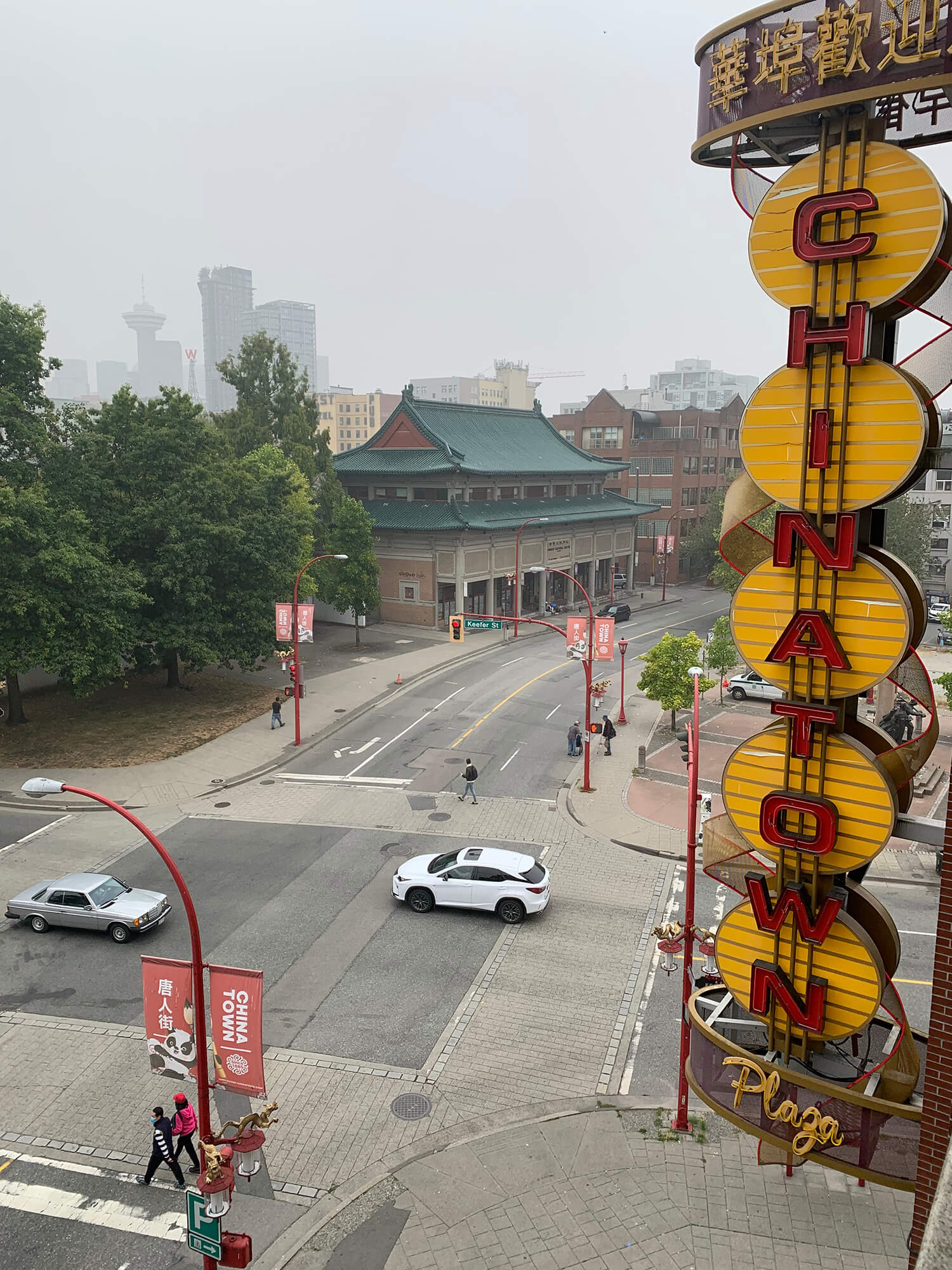Photo of the Chinatown Plaza sign on the corner of Keefer Street and Quebec Street in Vancouver Chinatown taken on an overcast day when the skyline is hazy with fog. The Chinese Cultural Centre Museum with its green roof is visible in the distance.