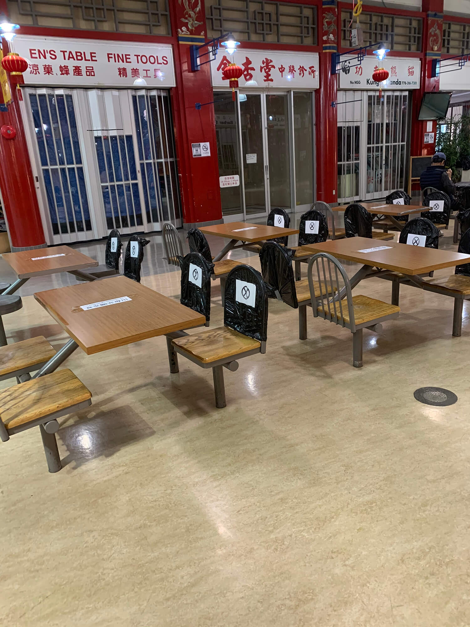 Empty food court tables inside Chinatown Plaza against a backdrop of dark and empty shop stalls with their gates and doors closed. Each table, which is intended to seat four, has three chairs marked as unavailable to encourage social distancing.