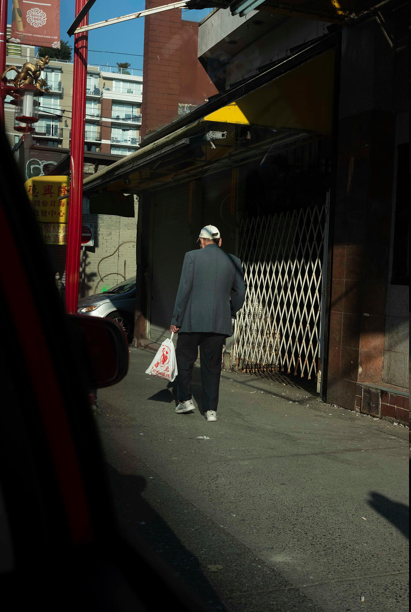 Image of an elderly man on a side street in Chinatown. He walks away from the camera as the sunlight falls across his back and lands on the closed and neglected storefronts he passes.