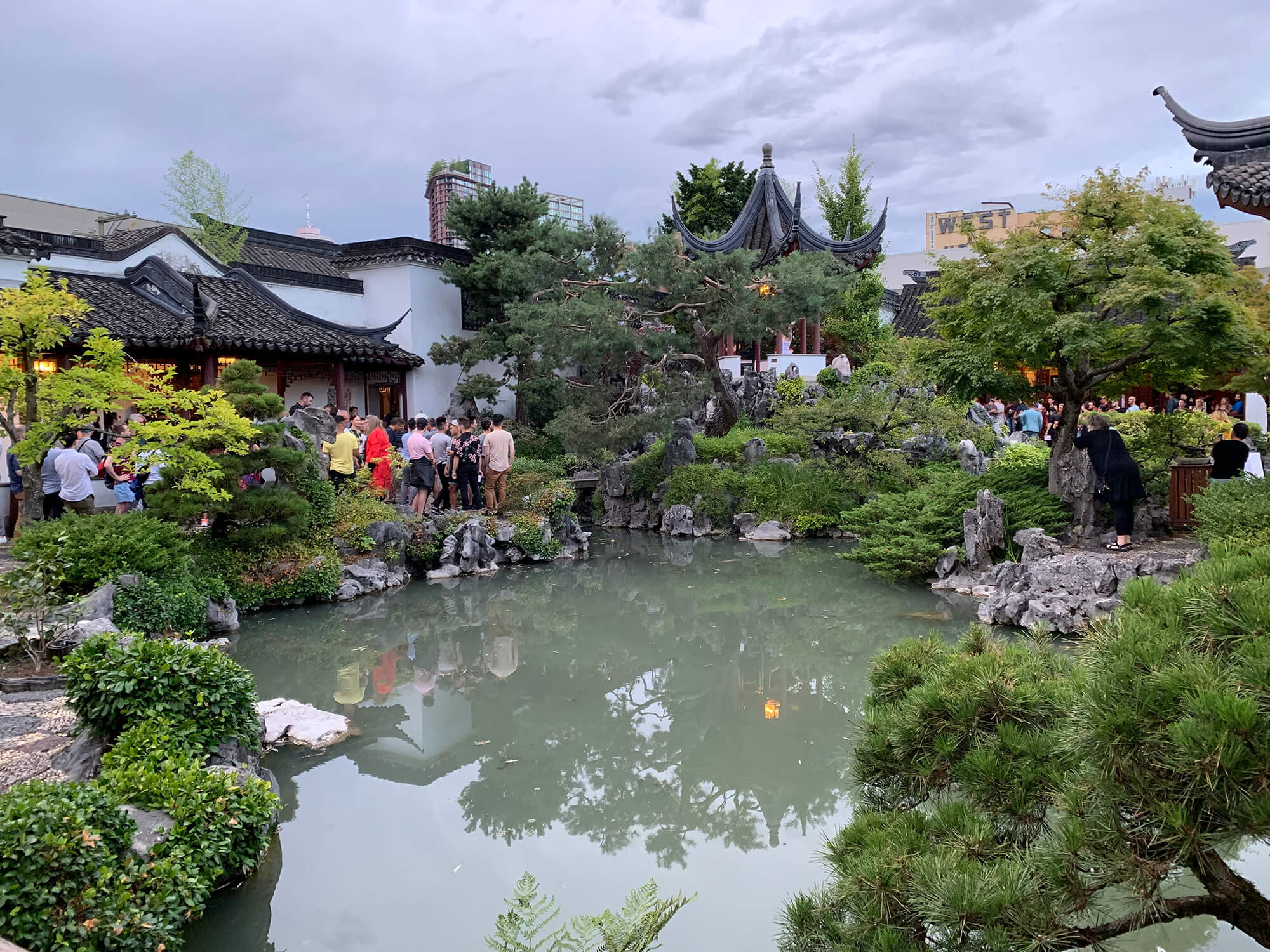Photo taken from inside Dr. Sun Yat-Sen Garden. The koi pond is lined with trees in green foliage. Across the water, crowds gather in the garden for Pride in Chinatown. It is dusk, and the light in the pavilion is reflected in the pond.