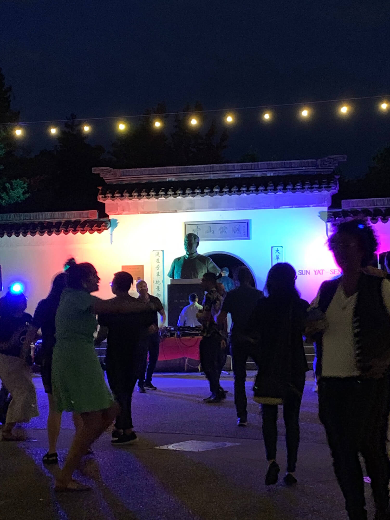 The courtyard between the Chinese Cultural Centre and Dr. Sun Yat-Sen Garden at night. The white garden wallis lit up in colours of the rainbow, while people dance in the foreground.