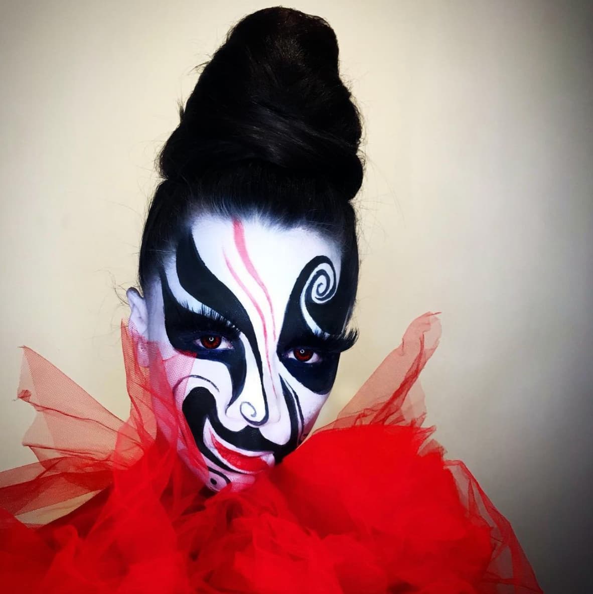 Maiden China in drag. Her long, black hair is pulled into a large hive that sits on top of her head. Her face is intricately painted in the style of Peking opera, but done so asymmetrically and with hints of abstractness. A red sheer ruffle collar obscures her chin.