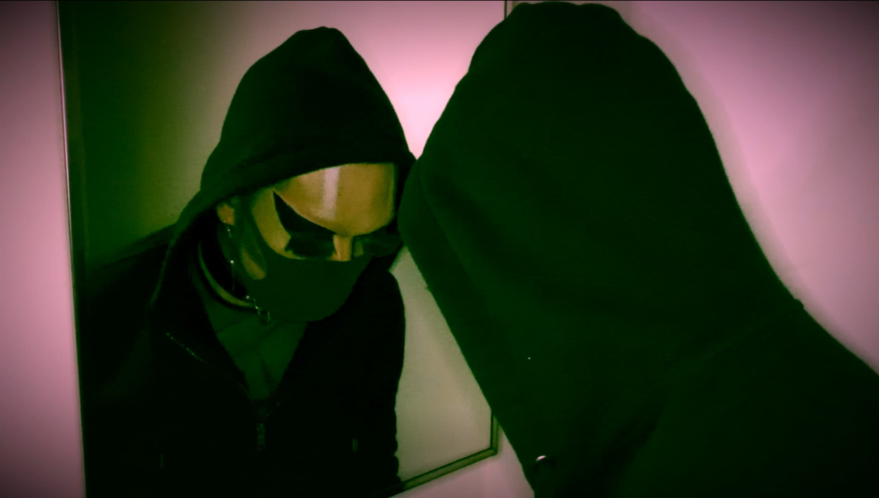 A still of Shay Dior in drag from their piece titled 'Contaminated by Racism'. Shay faces away from the camera, but we see their face reflected in the mirror in front of them. They have on a black cloth mask. The room is dark. We see Shay with their eyes downcast, which emphasizes their dramatic eye makeup and eyelash extensions.