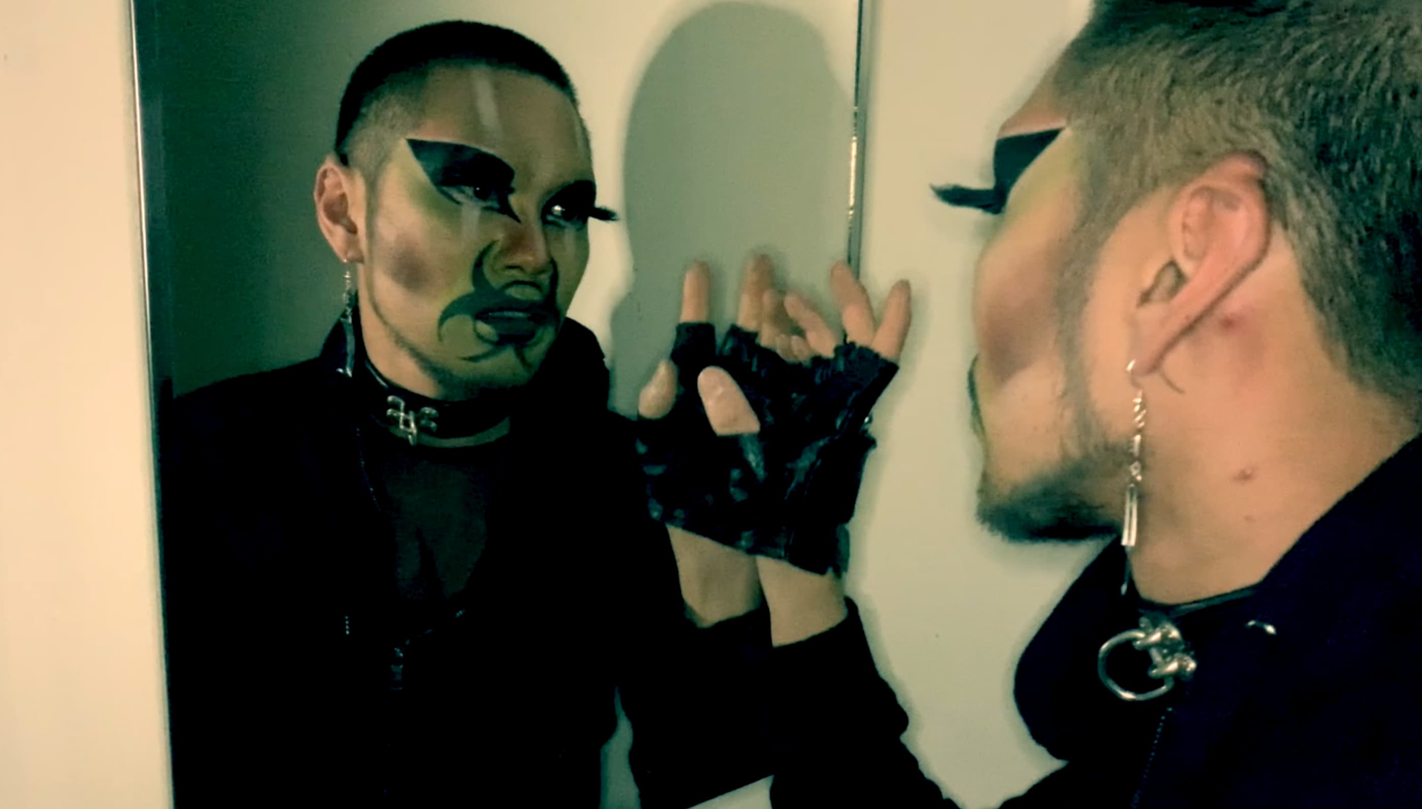 Still of Shay from 'Contaminated by Racism'. Shay has a closely cropped head, they're wearing a black leather choker, and a silver earring dangles from their left ear. They have a black biohazard symbol painted over their mouth. They look at their reflection in the mirror, and gently places their right hand on the glass surface.