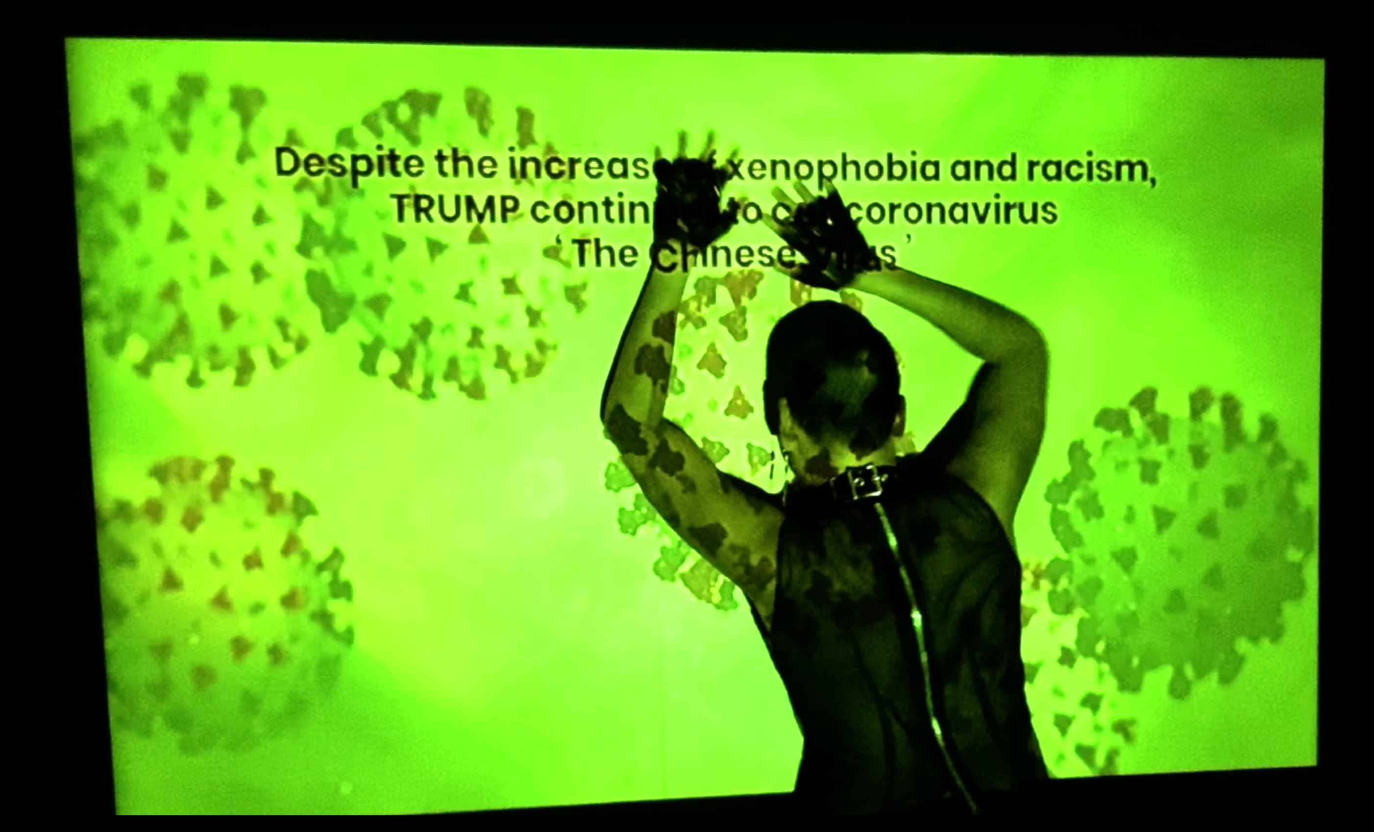 Still from 'Contaminated by Racism'. Shay faces away from the camera, they stand with their hands against a screen, onto which is projected representations of coronavirus particles in a greenish yellow tinge. Shay tries to cover overhead text that reads, ‘Despite the increase of xenophobia nad racism, TRUMP continues to call coronavirus “The Chinese virus”’.