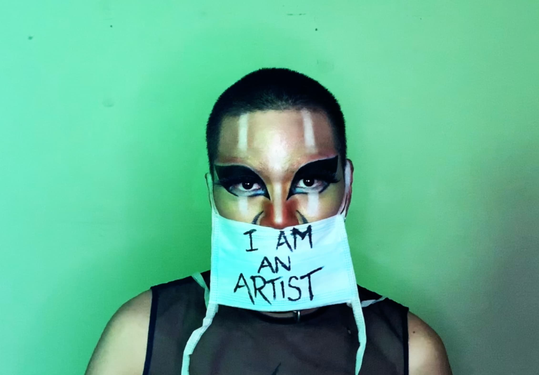 Still from 'Contaminated by Racism'. Shay stands against a green background. A blue medical mask covers their face below the nose, and it reads, ‘I am an artist’.