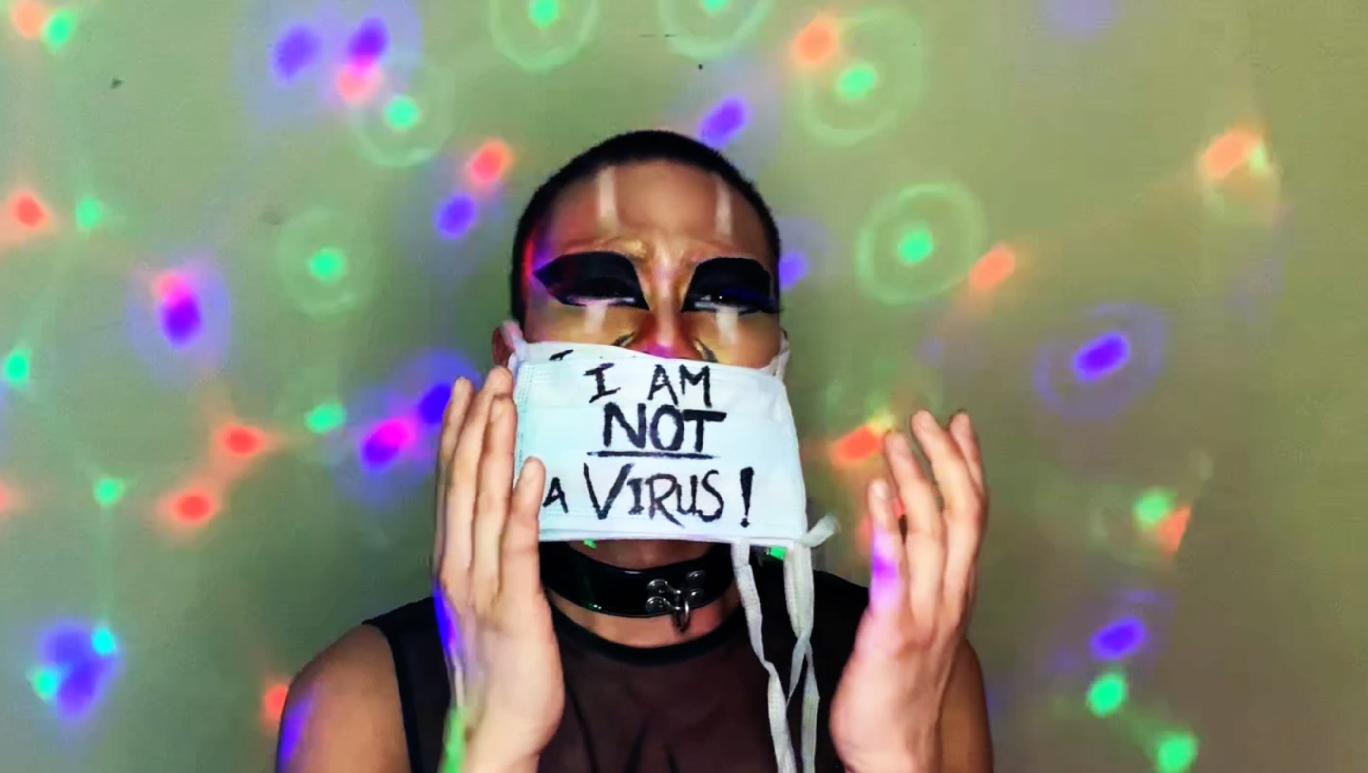 Still from 'Contaminated by Racism'. Shay stands against the same green background, now covered in projected moving lights in green, red, pink, and purple. They are wearing multiple medical masks, the one on top reads, ‘I am not a virus!’