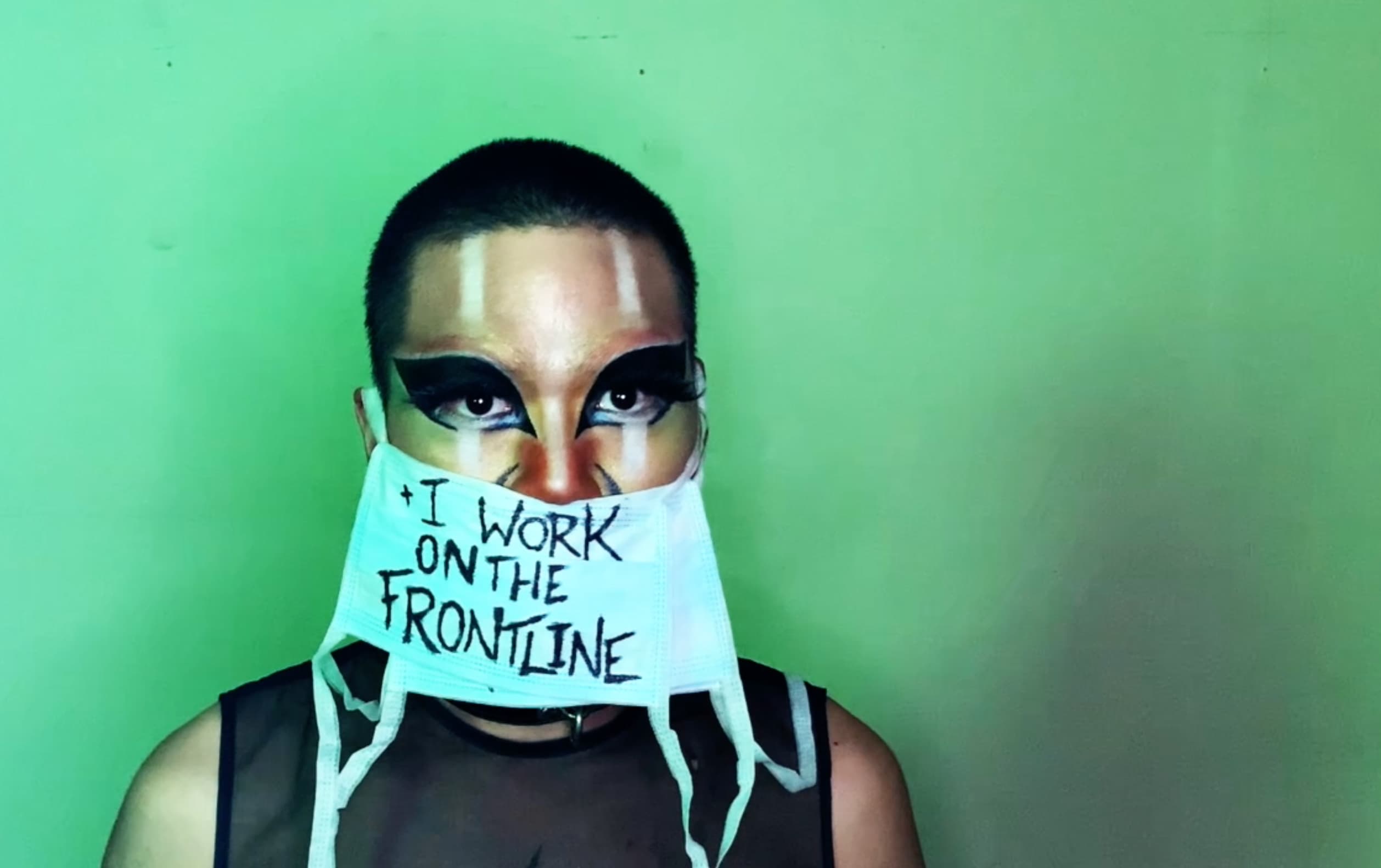 Still from 'Contaminated by Racism'. Shay stands against the same green background, wearing multiple medical masks. The one on top reads, ‘and I work on the frontline’.