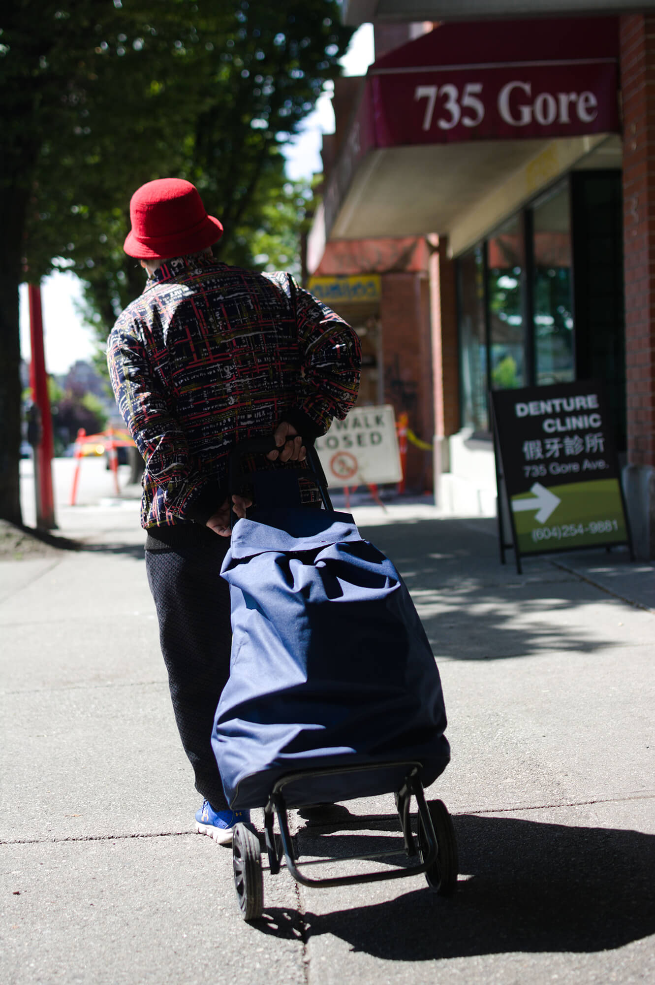 A Chinese senior in a red bowler hat walks away from the camera along Gore Street in Chinatown with a blue shopping bag in tow. The sunlight falls across her left shoulder and casts a shadow on the sidewalk. In front of her, out of focus, are pylons and a large white sign that announces the sidwalk is closed.