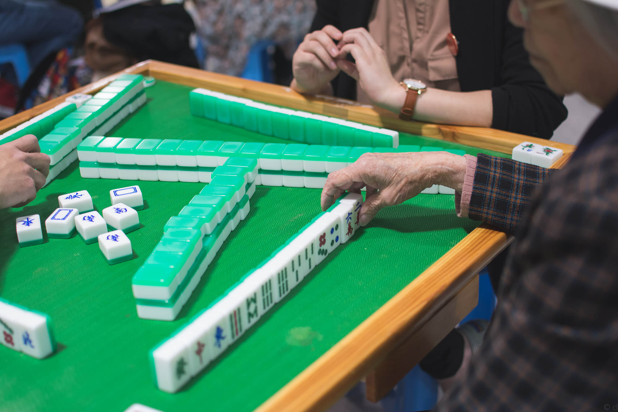 Youths and seniors play a round of mahjong. An elder has lined up her tiles neatly. She places her hand on a tile on the far right, perhaps in anticipation of making a move. This writer wishes they knew more about mahjong so they would know whether this is a good hand.