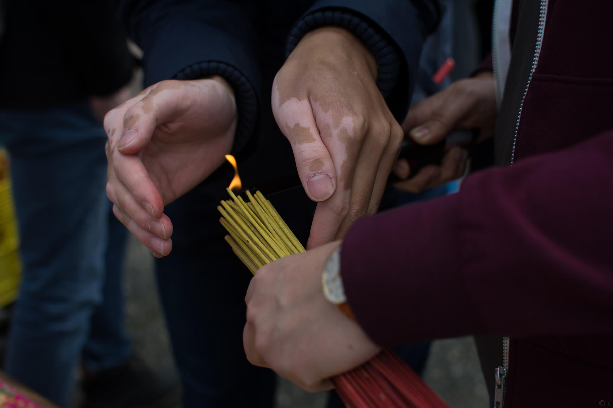 Close up of the hands of two people as they work together to light up joss sticks. One of them holds the bundle of yellow joss sticks in their left hand, while the other person cups their hands to protect the flame.