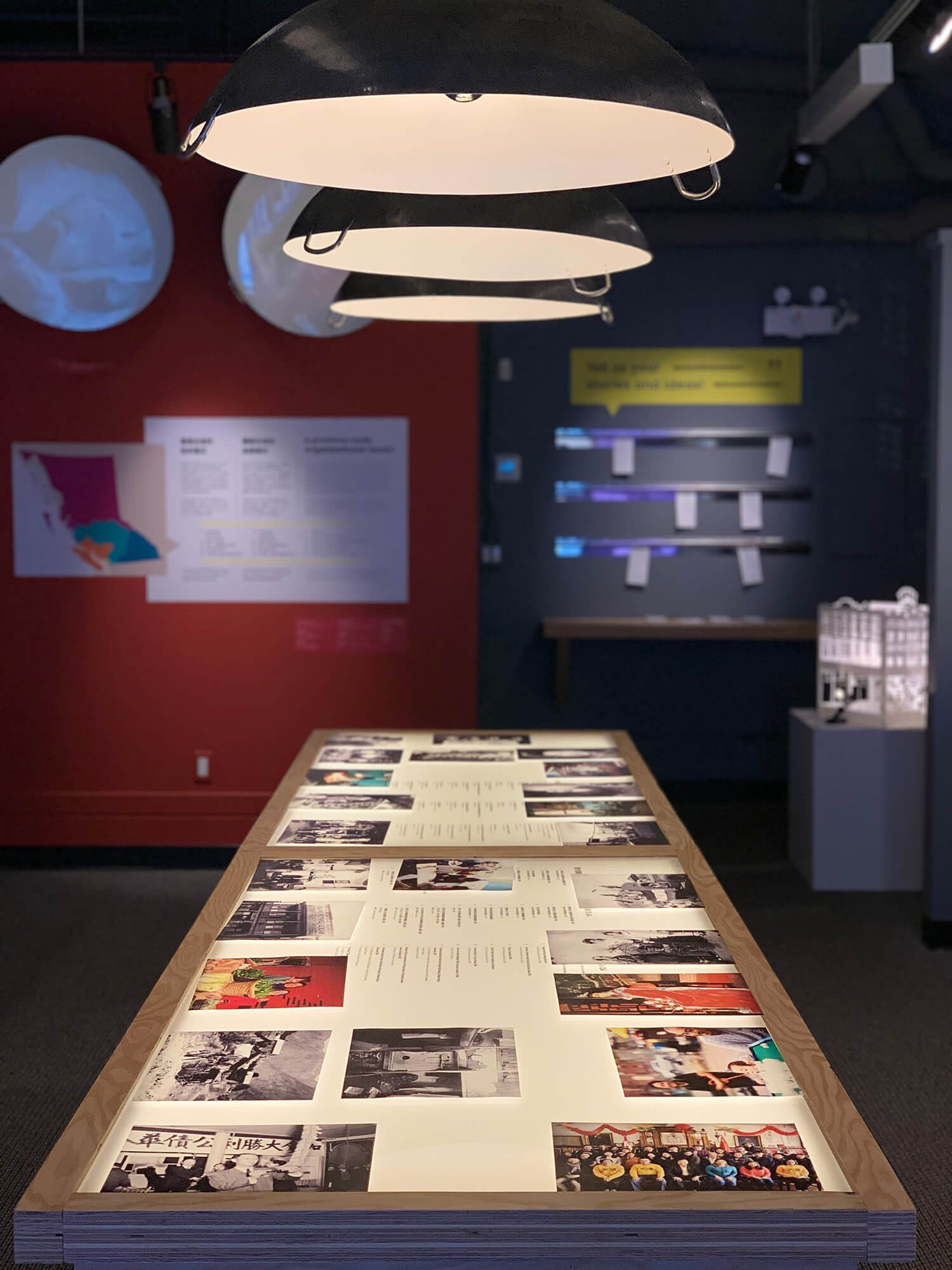 Interior of the Chinese Canadian Museum of British Columbia. A long display table covered in photographs is lit from above in lamps made out of woks.
