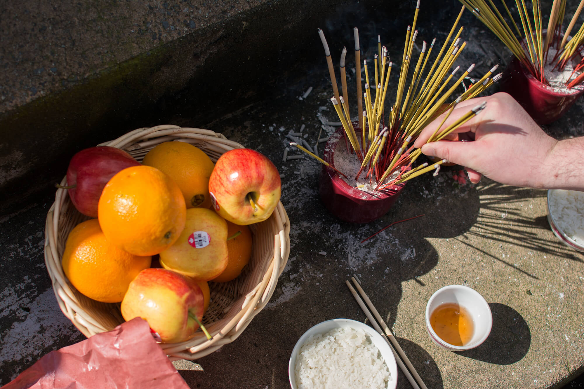 An ancestral offering of oranges and apples in a woven basket, a bowl of rice, a cup of tea, and two bowls of burning joss sticks. A hand places additional joss sticks into the incense bowl.