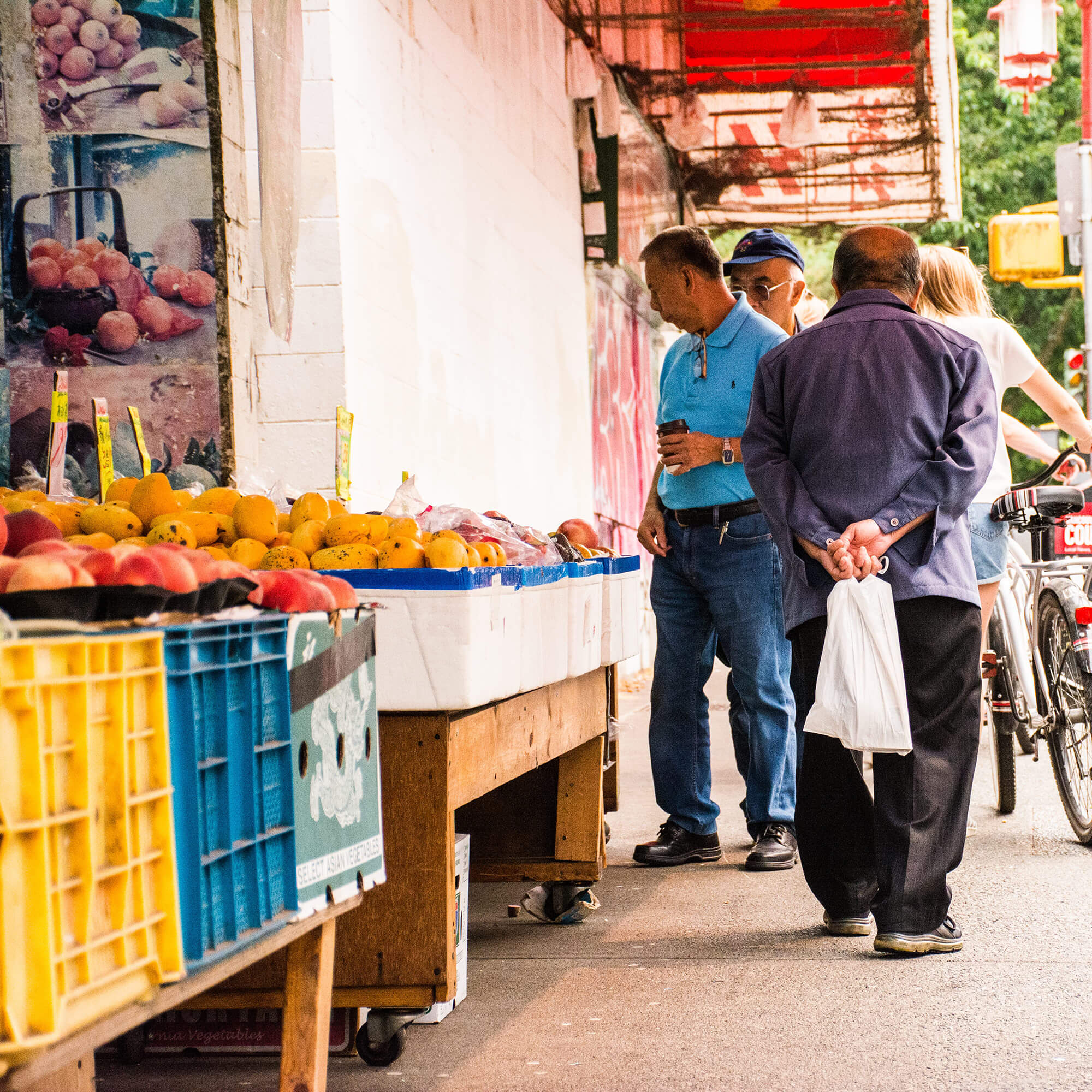 The sidewalk in front of a greengrocer in Chinatown. Wooden shelves on wheels carry plastic crates and styrofoam boxes overflowing with peaches, apples, mangos, and produce in pre-packaged bags. Passersby contemplate the fruits.