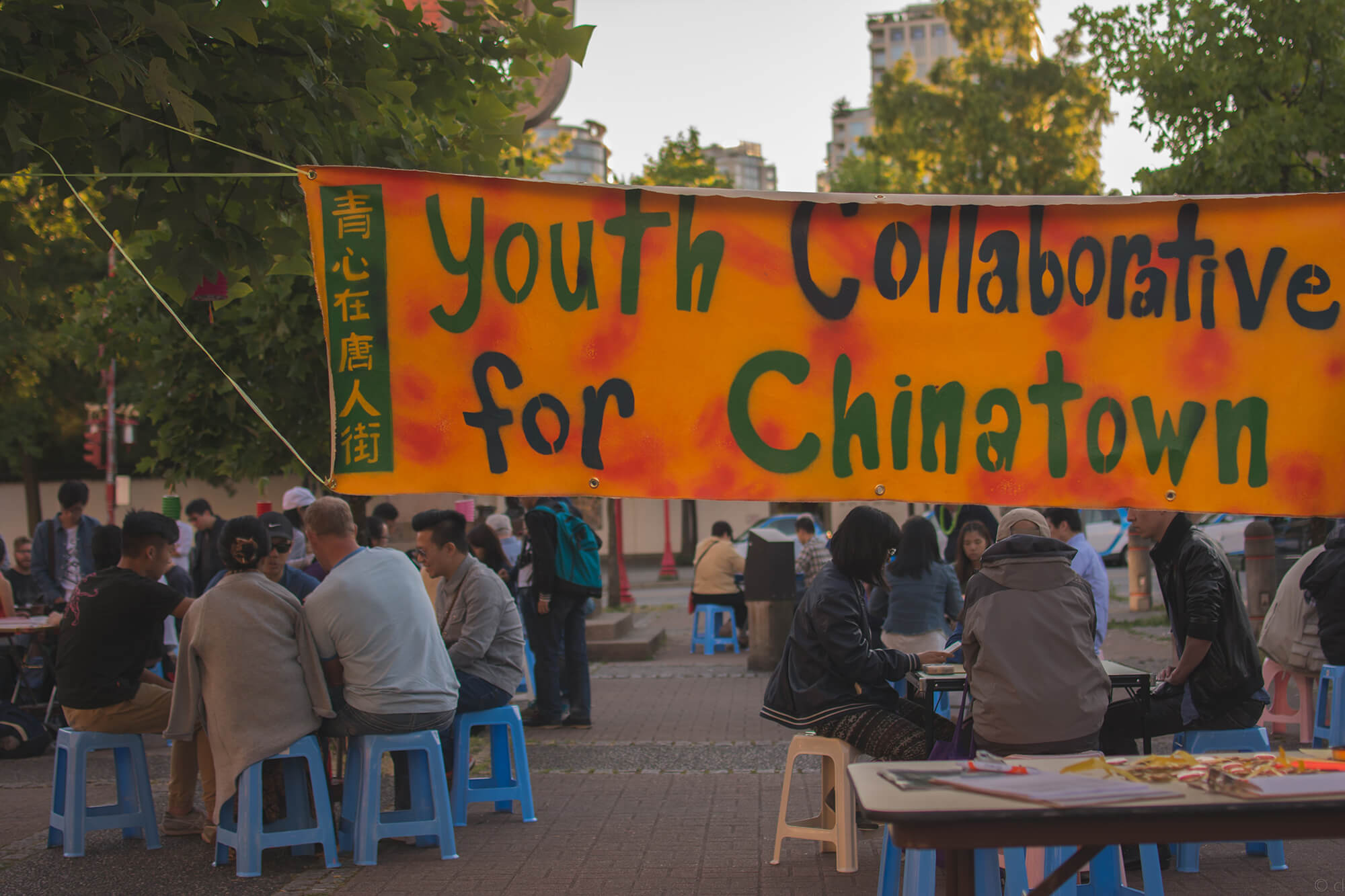 A Chinatown community event. Folks of all ages gather at dusk around tables in Chinatown Memorial Plaza, socializing, making crafts, and playing games. An orange banner with green text in the foreground reads, ‘Youth Collaborative for Chinatown’ and <span lang='zh'>青心在唐人街</span>.