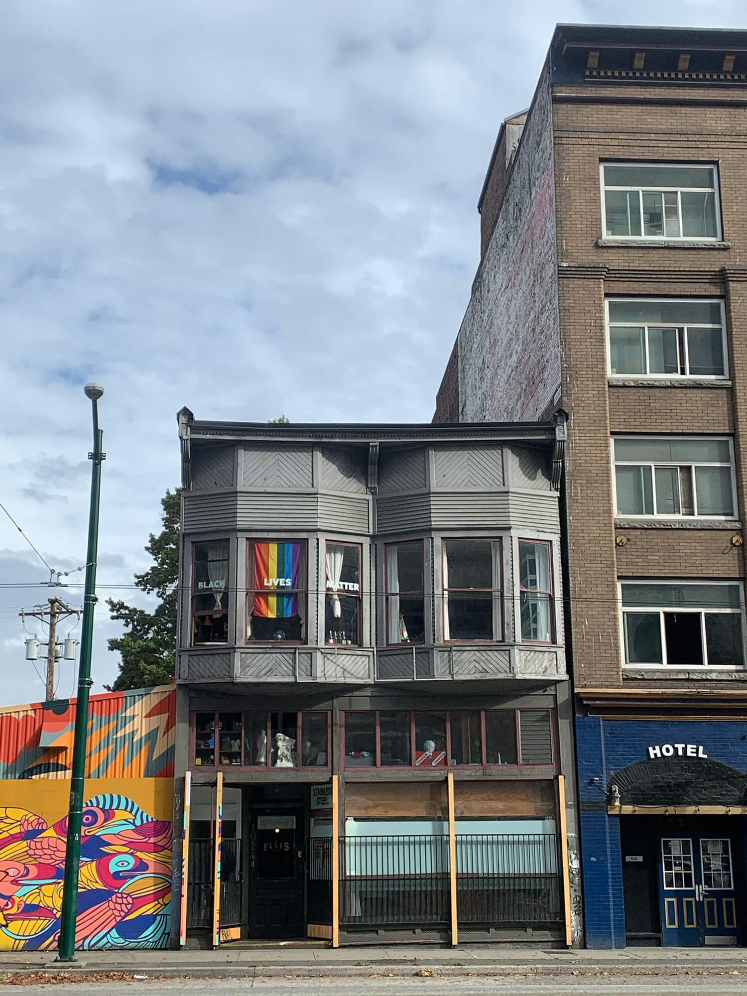 A gray two-storey walkup with two French windows on the second floor, flanked by a colourful mural of birds on the left and a hotel on the right. A raindow Pride flag hangs vertically from the French window on the left, behind white text that reads ‘Black Lives Matter’.