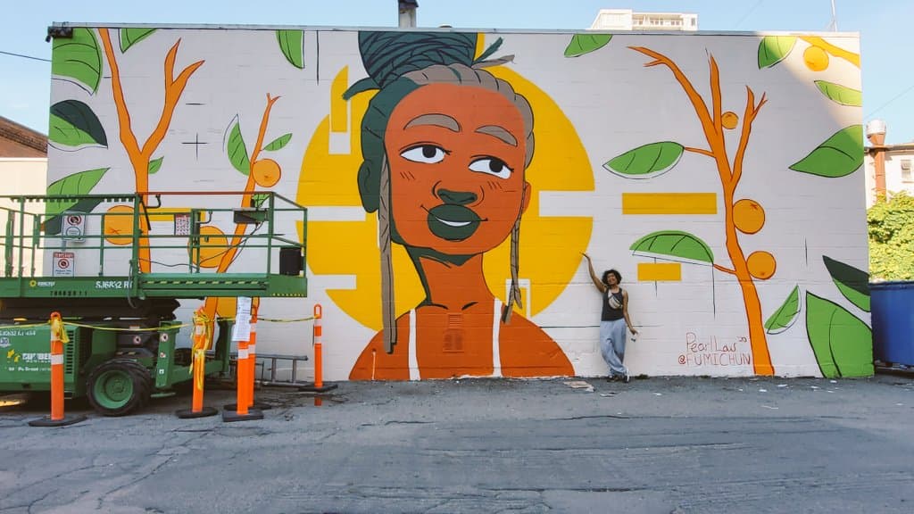 Pearl Low poses in front of her mural titled Precious Fruit. In the centre of the mural is a Black person with their braids pulled up into a bun while loose strands near their temples frame their face. They smile softly against the backdrop of a yellow circle, representing the sun. They are flanked by branches of orange trees, bearing ripe fruits and vibrant green leaves.