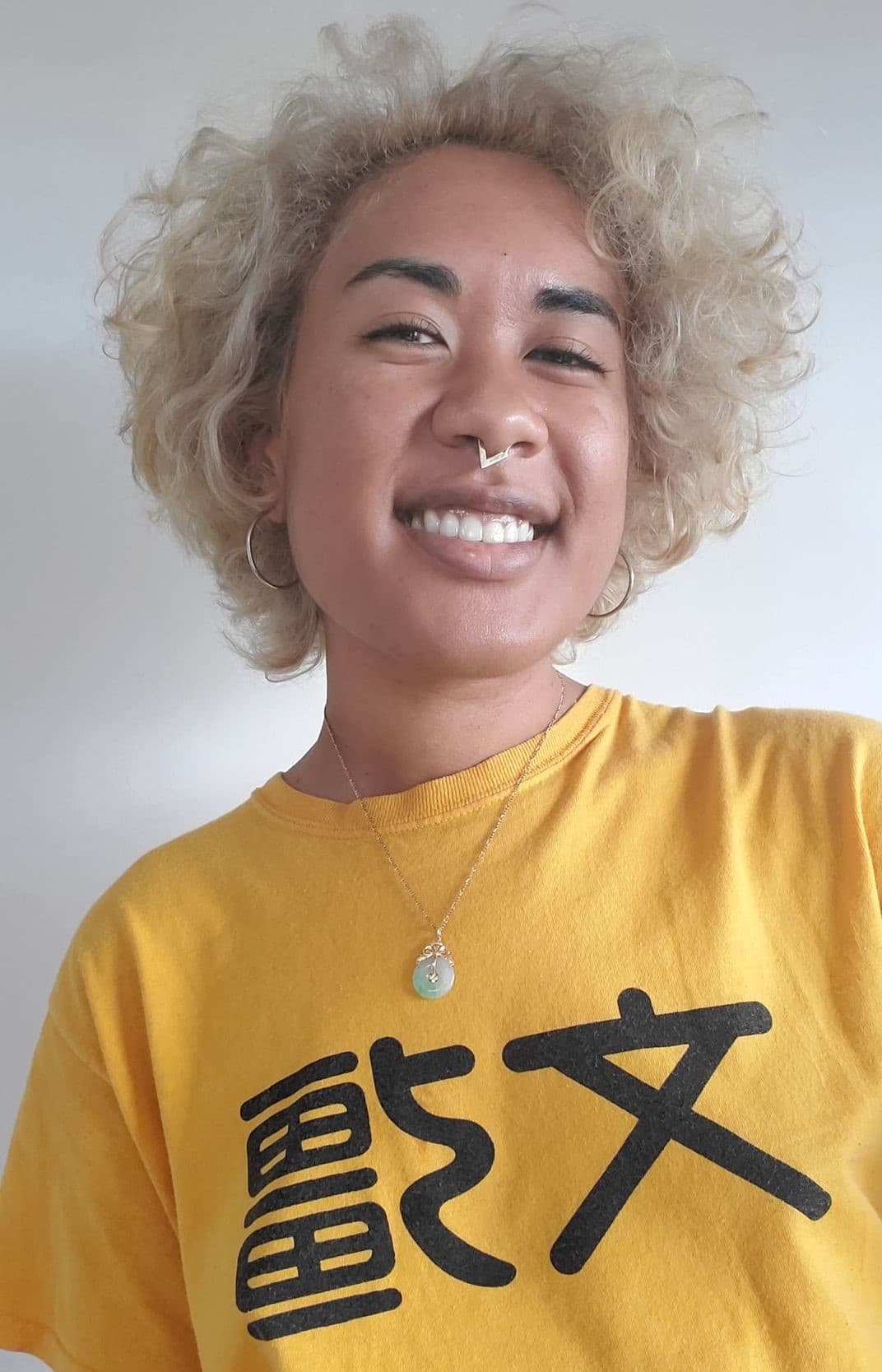 Pearl smiles brightly into the camera. This is a selfie. Her hair is dyed blonde and in loose curls, she wears hoop earrings, a septum piercing, a yellow t-shirt from Cantonese school, and a small, round jade pendant around her neck.